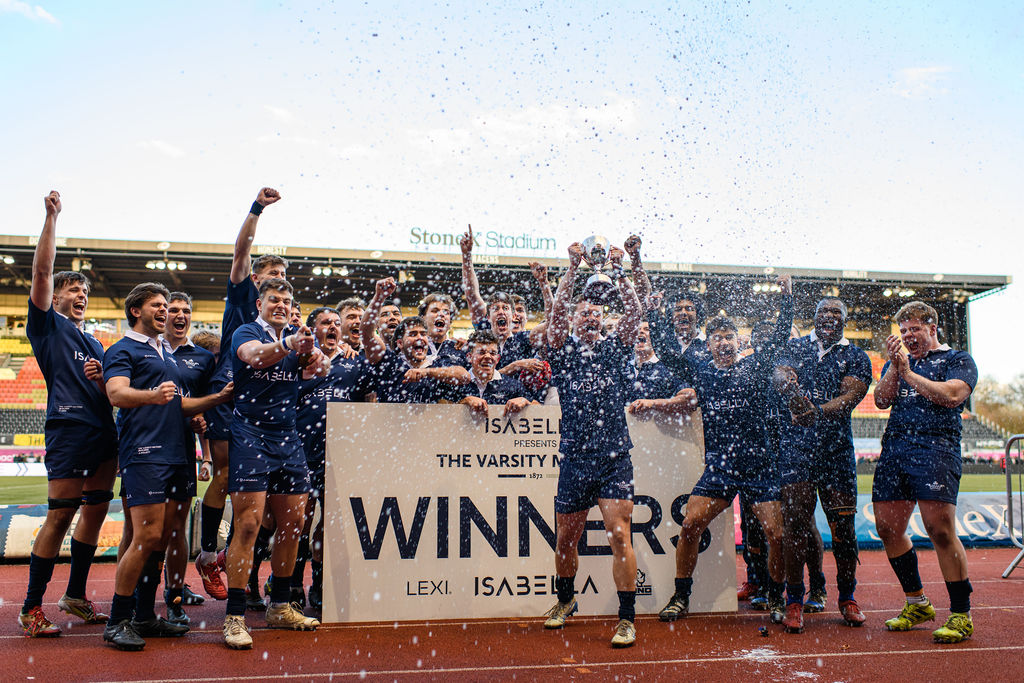 Oxford’s Men’s Blues celebrate their Varsity victory at StoneX Stadium, lifting the trophy behind a winners’ board as water sprays into the air. The team, dressed in navy kits, cheer with arms raised on the pitch following their win over Cambridge.